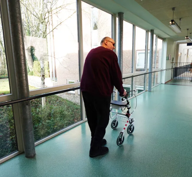 Bewoner Het Schild loopt in de gang met trillende rollator